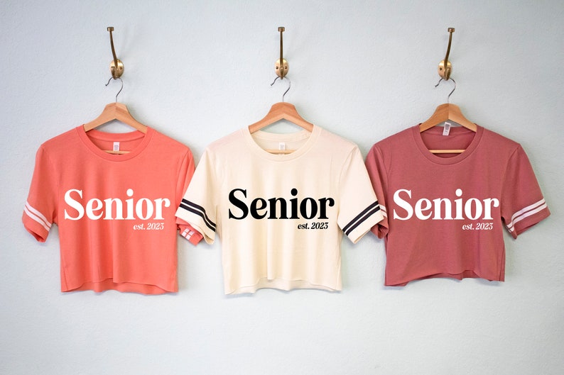 May include: Three cropped t-shirts with the word "Senior" and the text "est. 2023" on them. The shirts are in coral, cream, and dusty rose.