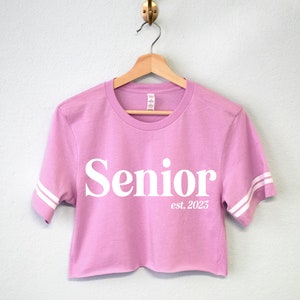 May include: A pink cropped t-shirt with white sleeves and the text "Senior est. 2023" printed on the front.