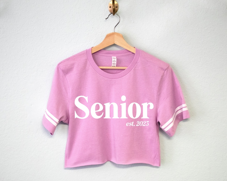 May include: A pink cropped t-shirt with white sleeves and the text "Senior est. 2023" printed on the front.