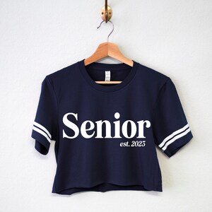 May include: A navy blue cropped t-shirt with white stripes on the sleeves and the words "Senior est. 2025" printed in white on the front.