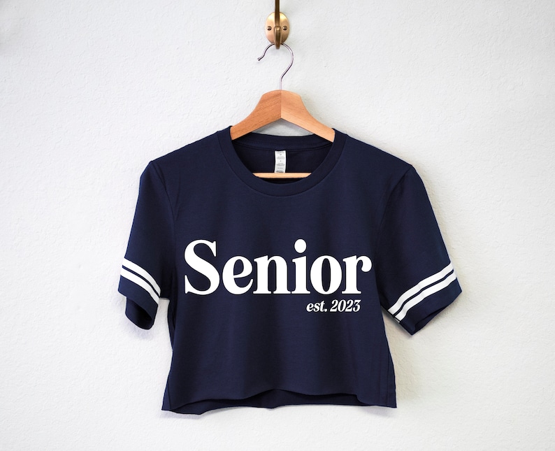 May include: A navy blue cropped t-shirt with white stripes on the sleeves and the words "Senior est. 2025" printed in white on the front.