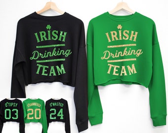 Puede incluir: Dos sudaderas cortas de color negro y verde con el texto "Irish Drinking Team" en purpurina dorada. Las sudaderas tienen diferentes números en la parte posterior: 03, 20 y 24.