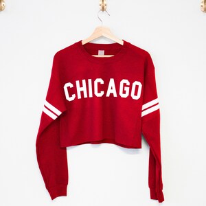 以下が含まれることがあります： 赤い長袖の白いクロップドスウェットシャツで、前面に白い「CHICAGO」の文字がプリントされています。