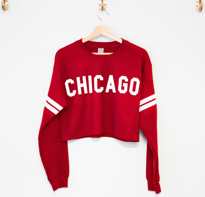 以下が含まれることがあります： 赤い長袖の白いクロップドスウェットシャツで、前面に白い「CHICAGO」の文字がプリントされています。