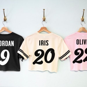 K&ouml;nnte beinhalten: Drei verk&uuml;rzte Trikots mit schwarzer und wei&szlig;er Beschriftung. Das schwarze Trikot tr&auml;gt die Aufschrift "JORDAN 9", das cremefarbene Trikot "IRIS 20" und das rosafarbene Trikot "OLIVIA 22".