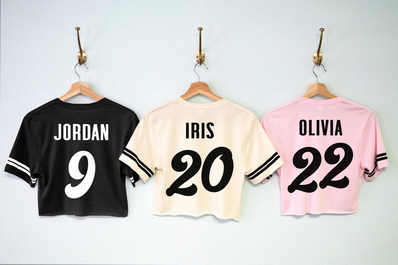 K&ouml;nnte beinhalten: Drei verk&uuml;rzte Trikots mit schwarzer und wei&szlig;er Beschriftung. Das schwarze Trikot tr&auml;gt die Aufschrift "JORDAN 9", das cremefarbene Trikot "IRIS 20" und das rosafarbene Trikot "OLIVIA 22".