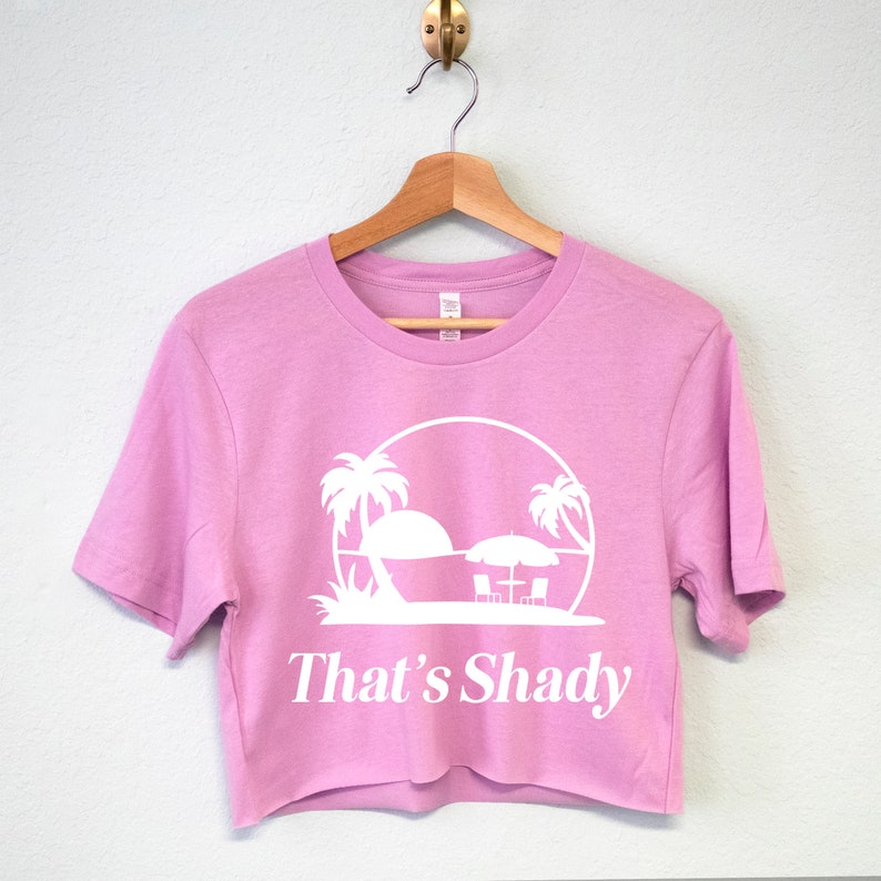 Puede incluir: Una camiseta rosa recortada con un gr&aacute;fico blanco de palmeras, una puesta de sol y dos tumbonas bajo una sombrilla. El texto "That's Shady" est&aacute; impreso debajo del gr&aacute;fico.