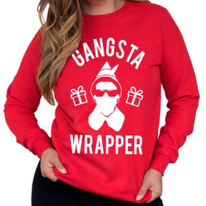May include: Red Christmas sweater with a white graphic of an elf wearing sunglasses and a face mask. The text "Gangsta Wrapper" is printed on the sweater.
