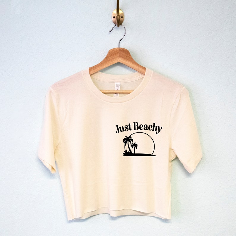 May include: A cream-colored cropped t-shirt with short sleeves and a black graphic of palm trees and a sun with the text "Just Beachy".