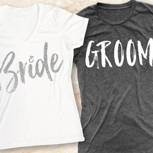 K&ouml;nnte beinhalten: Ein wei&szlig;es T-Shirt mit dem Wort "Bride" in silbernen Glitzerbuchstaben und ein graues T-Shirt mit dem Wort "Groom" in wei&szlig;en Buchstaben.