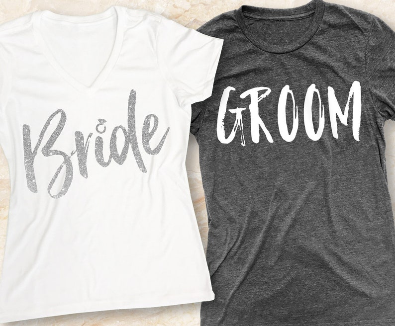 K&ouml;nnte beinhalten: Ein wei&szlig;es T-Shirt mit dem Wort "Bride" in silbernen Glitzerbuchstaben und ein graues T-Shirt mit dem Wort "Groom" in wei&szlig;en Buchstaben.