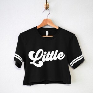 K&ouml;nnte beinhalten: Schwarzes, bauchfreies T-Shirt mit wei&szlig;en Streifen an den &Auml;rmeln und dem Wort "Little" in wei&szlig;er Retro-Schrift.