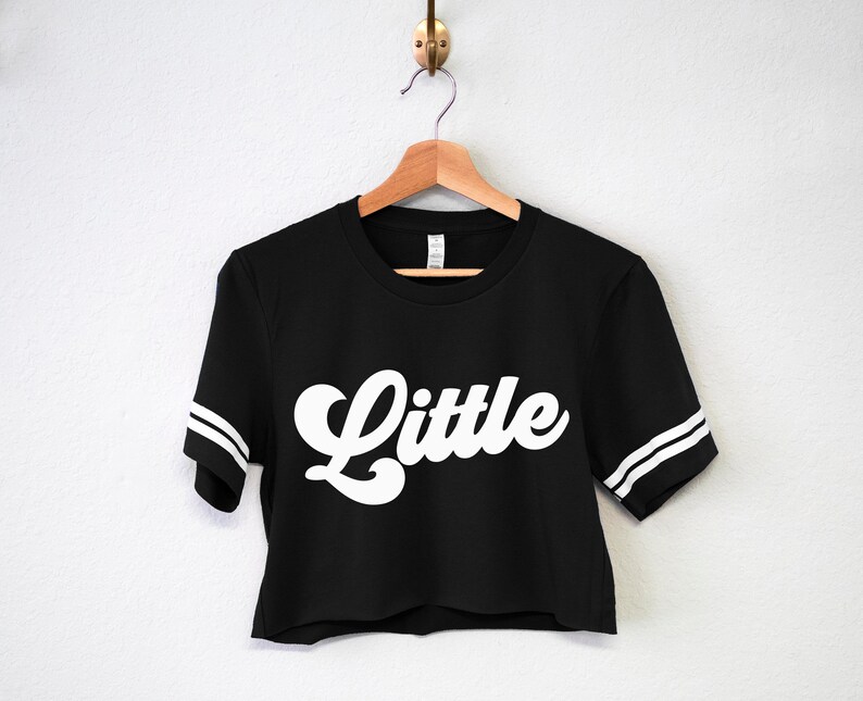 K&ouml;nnte beinhalten: Schwarzes, bauchfreies T-Shirt mit wei&szlig;en Streifen an den &Auml;rmeln und dem Wort "Little" in wei&szlig;er Retro-Schrift.
