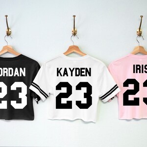 May include: Three cropped t-shirts in black, white and pink with black and white striped sleeves. The shirts have the number 23 and a name printed on the back. The names are Jordan, Kayden, and Iris.