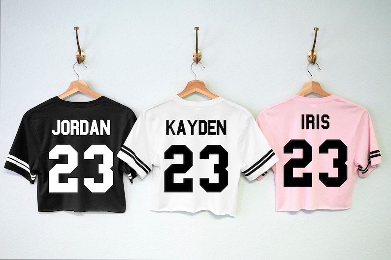 May include: Three cropped t-shirts in black, white and pink with black and white striped sleeves. The shirts have the number 23 and a name printed on the back. The names are Jordan, Kayden, and Iris.