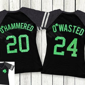 May include: Black raglan-sleeve shirts with green glitter text. One shirt says "O'HAMMERED 20" and the other says "O'WASTED 24". Each shirt has a small green shamrock on the front. The sleeves are dark grey with white stripes.