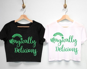 May include: Two cropped t-shirts with the text "magically delicious" in green glitter lettering. One shirt is black and the other is white. Both shirts feature a green glitter rainbow with a shamrock at the end.