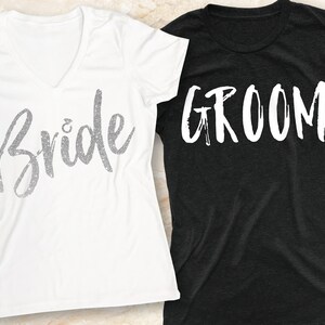 K&ouml;nnte beinhalten: Zwei T-Shirts, eines wei&szlig; und eines schwarz, mit den Worten "Bride" und "Groom" in wei&szlig;er, Pinselstrichschrift.