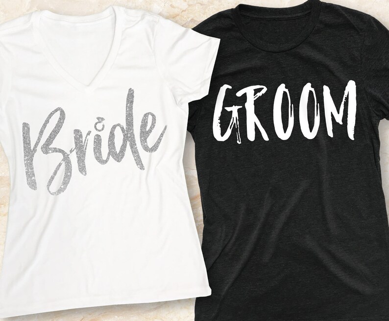 K&ouml;nnte beinhalten: Zwei T-Shirts, eines wei&szlig; und eines schwarz, mit den Worten "Bride" und "Groom" in wei&szlig;er, Pinselstrichschrift.
