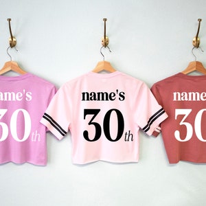 May include: Three pink and red cropped t-shirts with black and white striped sleeves. The shirts have "name's 30th" printed on the back.