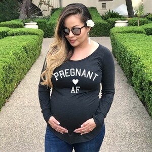 Puede incluir: Una mujer embarazada lleva una camiseta negra de manga larga con el texto "Pregnant AF" y un coraz&oacute;n blanco. Est&aacute; de pie en un jard&iacute;n con setos verdes.