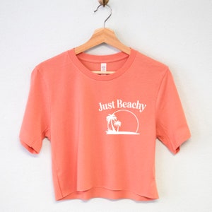 May include: A coral colored cropped t-shirt with short sleeves and a white graphic of palm trees and a sun with the text "Just Beachy" on the front.