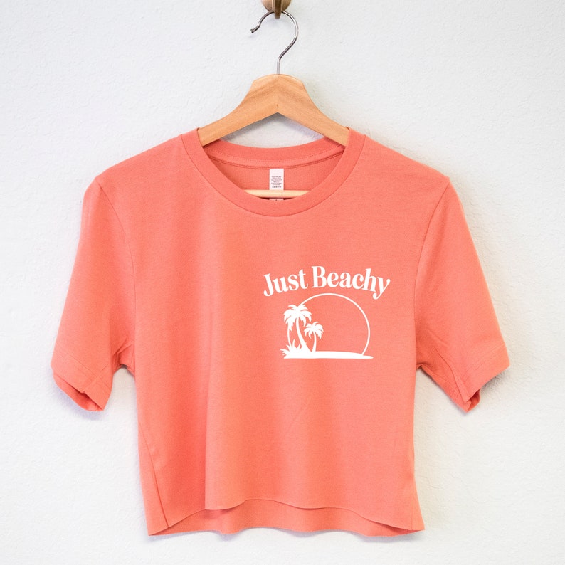 May include: A coral colored cropped t-shirt with short sleeves and a white graphic of palm trees and a sun with the text "Just Beachy" on the front.