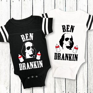 Op de afbeelding: Twee zwart-witte baby rompers met rode en witte strepen op de mouwen. De rompers hebben een zwart-witte illustratie van Benjamin Franklin met een zonnebril en twee babyflesjes. De tekst op de rompers is "BEN DRANKIN".