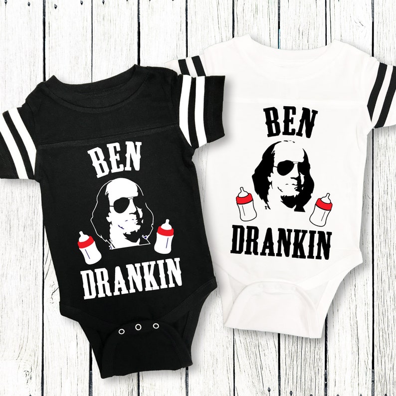 Op de afbeelding: Twee zwart-witte baby rompers met rode en witte strepen op de mouwen. De rompers hebben een zwart-witte illustratie van Benjamin Franklin met een zonnebril en twee babyflesjes. De tekst op de rompers is "BEN DRANKIN".