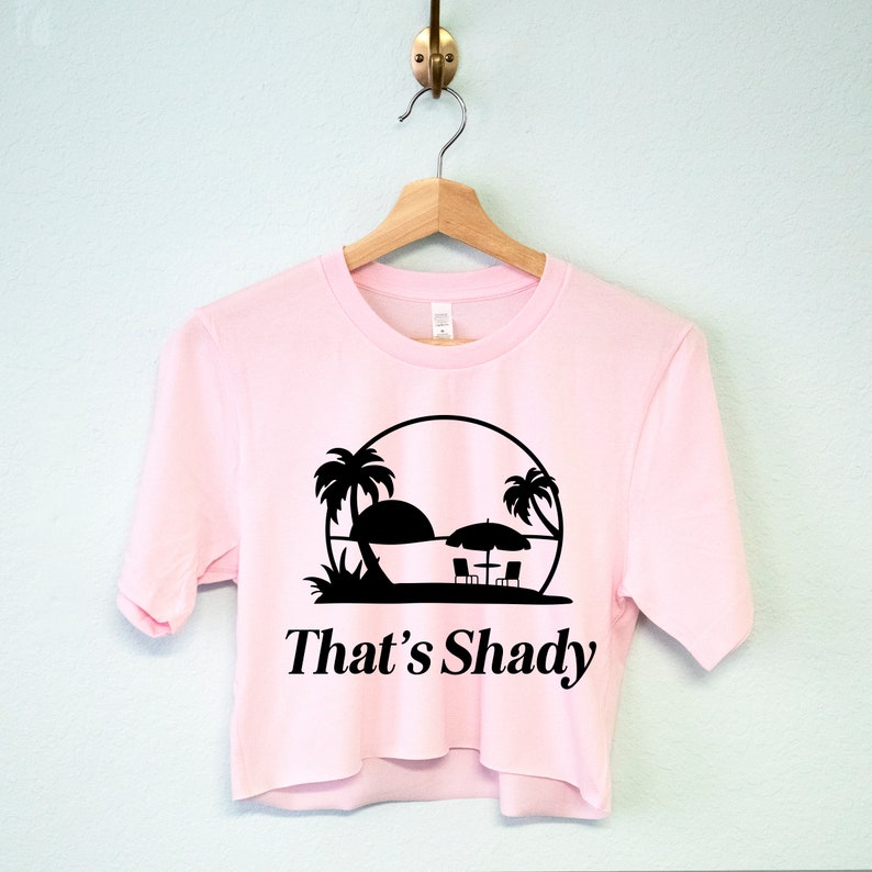 Puede incluir: Una camiseta rosa de manga corta y corte corto con un gr&aacute;fico negro de una silueta de palmera y el texto "That's Shady".