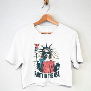 May include: White cropped t-shirt with a graphic of the Statue of Liberty holding a glass of wine and a passport. The text "Party in the USA" is printed below the graphic.