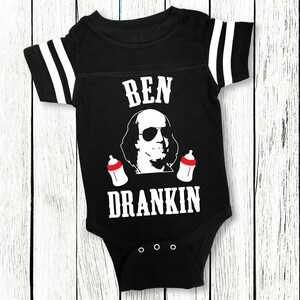 Op de afbeelding: Een zwart-witte baby romper met een voetbaltrui-ontwerp. Het heeft een cartoon afbeelding van Benjamin Franklin met een zonnebril en twee babyflesjes. De tekst "BEN DRANKIN" staat op de romper gedrukt.