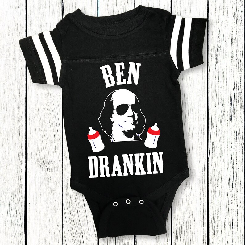 Op de afbeelding: Een zwart-witte baby romper met een voetbaltrui-ontwerp. Het heeft een cartoon afbeelding van Benjamin Franklin met een zonnebril en twee babyflesjes. De tekst "BEN DRANKIN" staat op de romper gedrukt.
