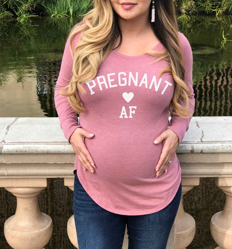 Puede incluir: Una camiseta rosa de manga larga con el texto "PREGNANT AF" en letras blancas con un coraz&oacute;n blanco entre las palabras.
