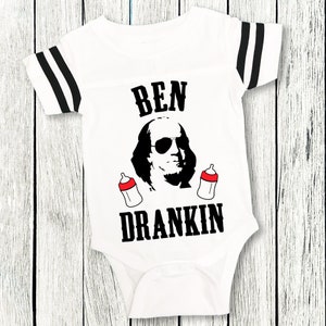 Op de afbeelding: Een witte baby romper met zwart-wit gestreepte mouwen. De romper heeft een zwart-witte afbeelding van Benjamin Franklin met een zonnebril en twee babyflesjes. De tekst "BEN DRANKIN" is in zwart onder de afbeelding gedrukt.