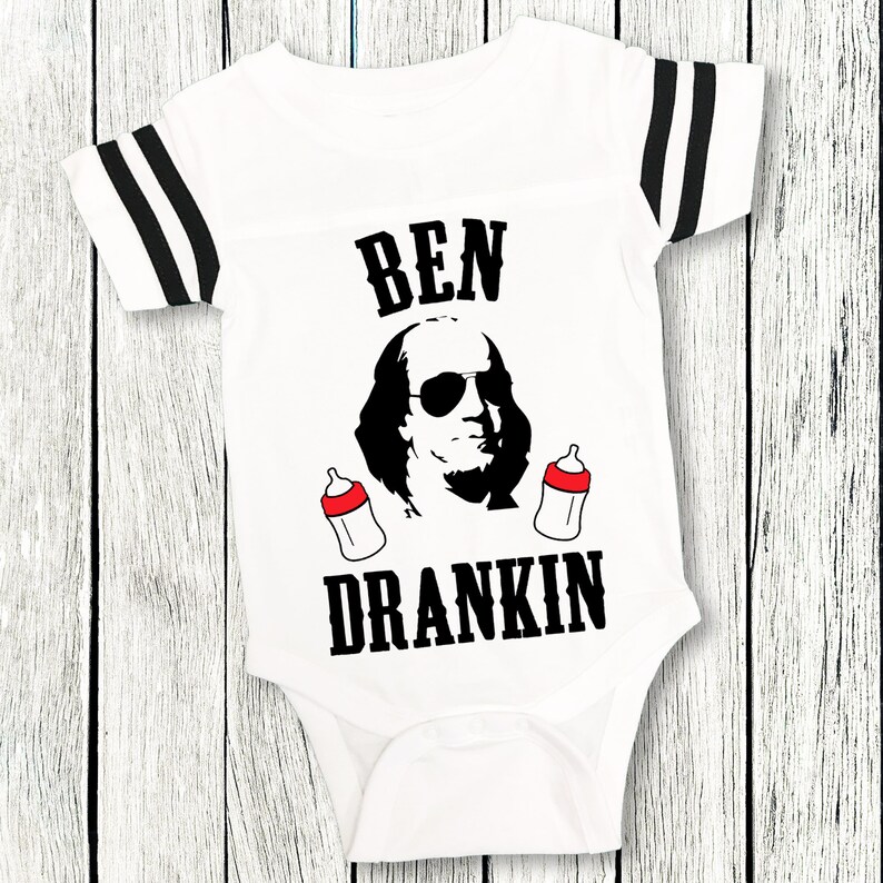 Op de afbeelding: Een witte baby romper met zwart-wit gestreepte mouwen. De romper heeft een zwart-witte afbeelding van Benjamin Franklin met een zonnebril en twee babyflesjes. De tekst "BEN DRANKIN" is in zwart onder de afbeelding gedrukt.