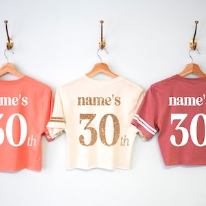 May include: Three cropped t-shirts with the text "name's 30th" on the back. The shirts are in coral, cream, and maroon.