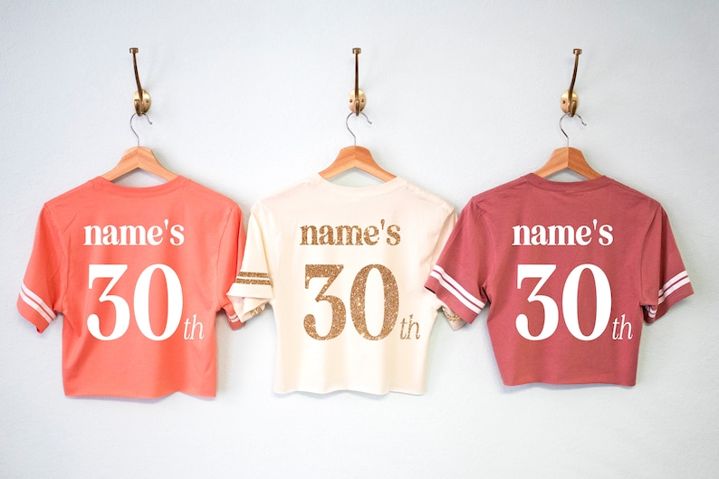 May include: Three cropped t-shirts with the text "name's 30th" on the back. The shirts are in coral, cream, and maroon.