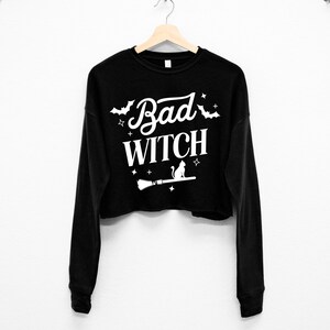 May include: Black cropped sweatshirt with long sleeves featuring a white graphic print of a cat sitting on a broomstick with the text "Bad Witch" and stars.