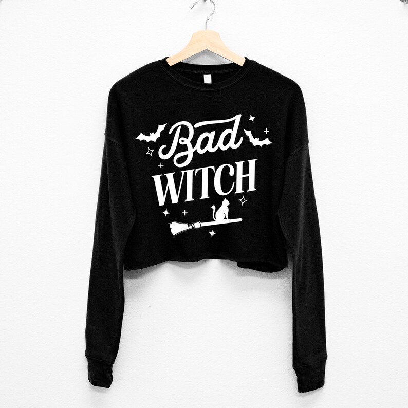 May include: Black cropped sweatshirt with long sleeves featuring a white graphic print of a cat sitting on a broomstick with the text "Bad Witch" and stars.