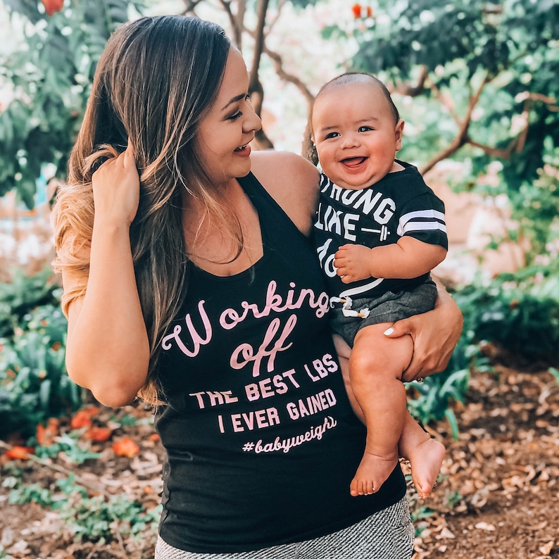 Pu&ograve; includere: Una donna indossa una canotta nera con la scritta "Working off the best lbs I ever gained #babyweight" in lettere rosa. Tiene in braccio un bambino che indossa una maglietta a righe bianche e nere con la scritta "Strong like 10" in lettere nere.