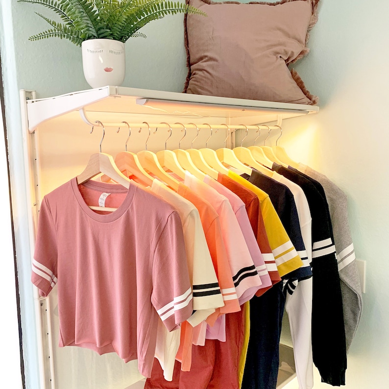 May include: A rack of clothing with a variety of colors and styles, including pink, yellow, blue, and white. The clothing is hanging on a white metal rack with wooden hangers. The rack is mounted on a white shelf.