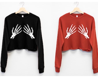 May include: Two cropped long-sleeved sweatshirts with a skeleton hand graphic. One sweatshirt is black and the other is red.
