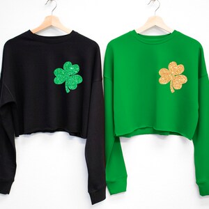 May include: Two cropped sweatshirts with long sleeves. One is black with a green glitter shamrock and the other is green with a gold glitter shamrock.