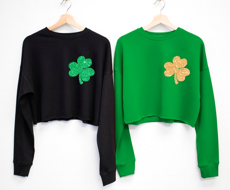 May include: Two cropped sweatshirts with long sleeves. One is black with a green glitter shamrock and the other is green with a gold glitter shamrock.
