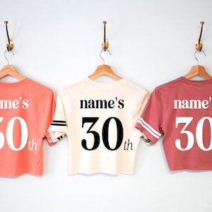 May include: Three cropped t-shirts in different colors, coral, cream, and rose, with the text "name's 30th" printed on the back.