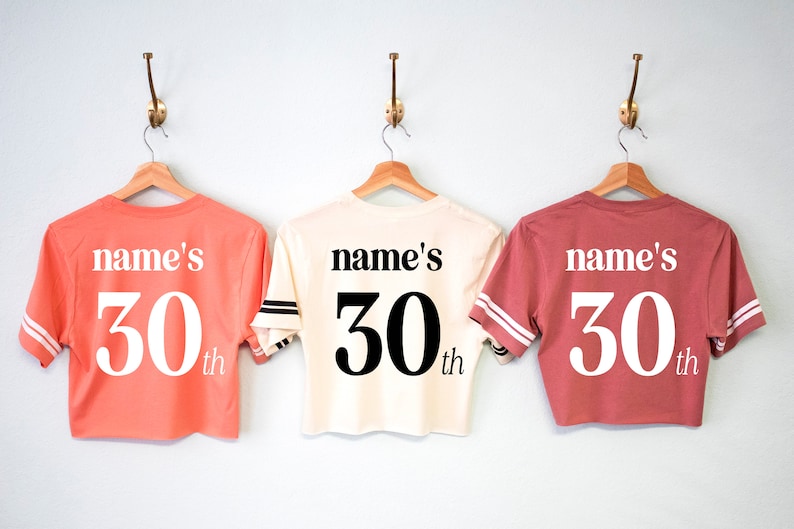 May include: Three cropped t-shirts in different colors, coral, cream, and rose, with the text "name's 30th" printed on the back.