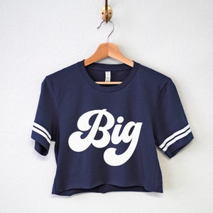 K&ouml;nnte beinhalten: Ein dunkelblaues, bauchfreies T-Shirt mit wei&szlig;en Streifen an den &Auml;rmeln und dem Wort "Big" in einer wei&szlig;en Retro-Schrift.