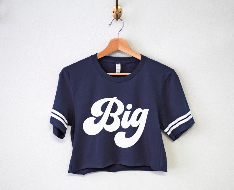 K&ouml;nnte beinhalten: Ein dunkelblaues, bauchfreies T-Shirt mit wei&szlig;en Streifen an den &Auml;rmeln und dem Wort "Big" in einer wei&szlig;en Retro-Schrift.