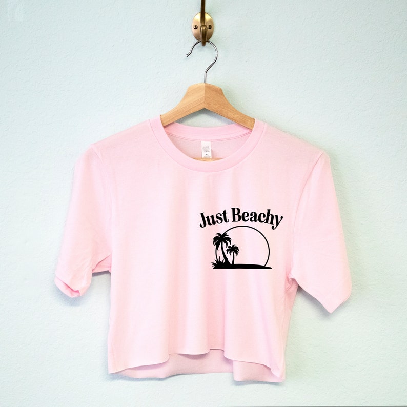 May include: A pink cropped t-shirt with short sleeves and a black graphic print of palm trees and a sun with the text "Just Beachy".